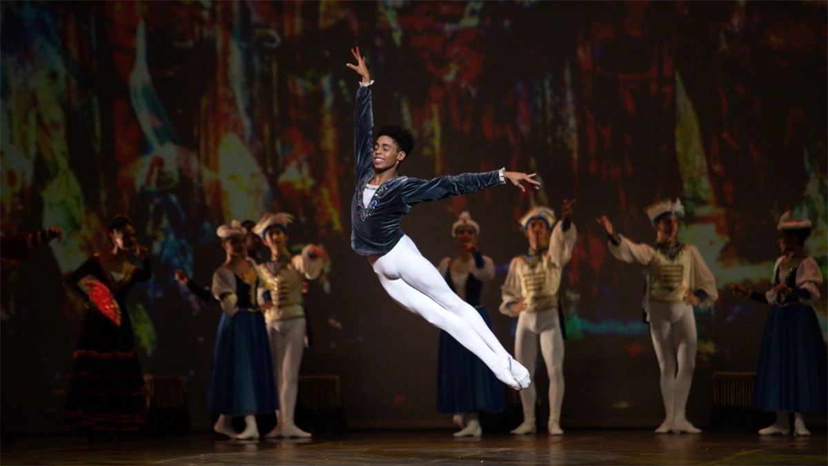A fotografia retrata o bailarino Enzo Santos, do Balé do Teatro Bolshoi no Brasil, em pleno salto durante uma apresentação. Ele veste collant branco e blusa azul escura adornada, com os braços elegantemente abertos e um sorriso no rosto, evidenciando leveza e técnica.