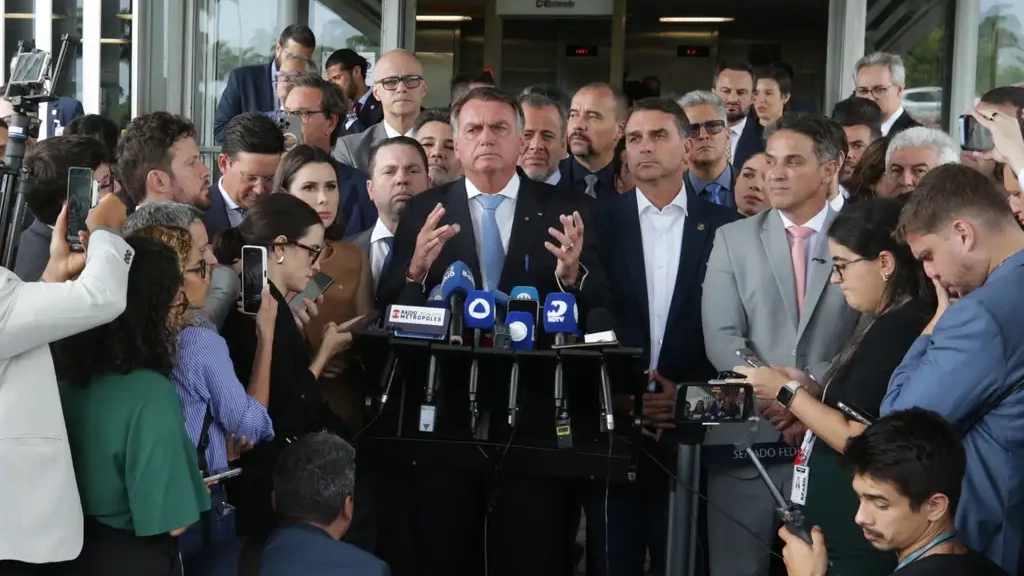 Ex-presidente Jair Bolsonaro faz pronunciamento à imprensa cercado por aliados e jornalistas em frente ao Congresso Nacional, em Brasília. Diversos microfones de veículos de mídia estão posicionados à sua frente enquanto ele gesticula com as mãos, em meio a grande cobertura da imprensa.