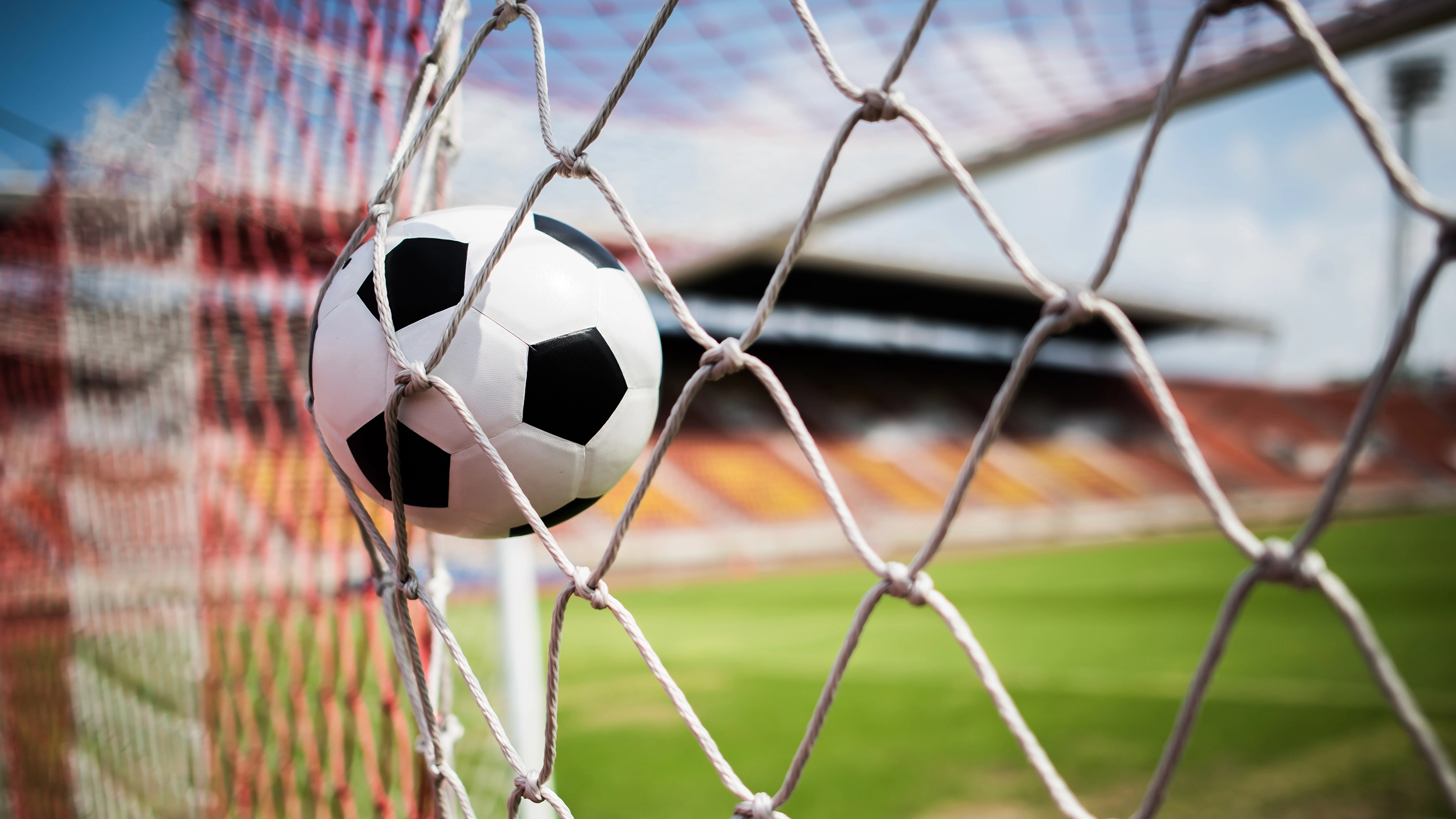A imagem mostra uma bola de futebol branca e preta presa na rede de um gol, indicando que acabou de ser marcada uma pontuação. O fundo exibe um estádio com arquibancadas vazias e céu azul, transmitindo uma atmosfera de jogo em campo aberto. A foto está em foco na bola e na rede, com o restante do estádio desfocado ao fundo, destacando o momento exato do gol.