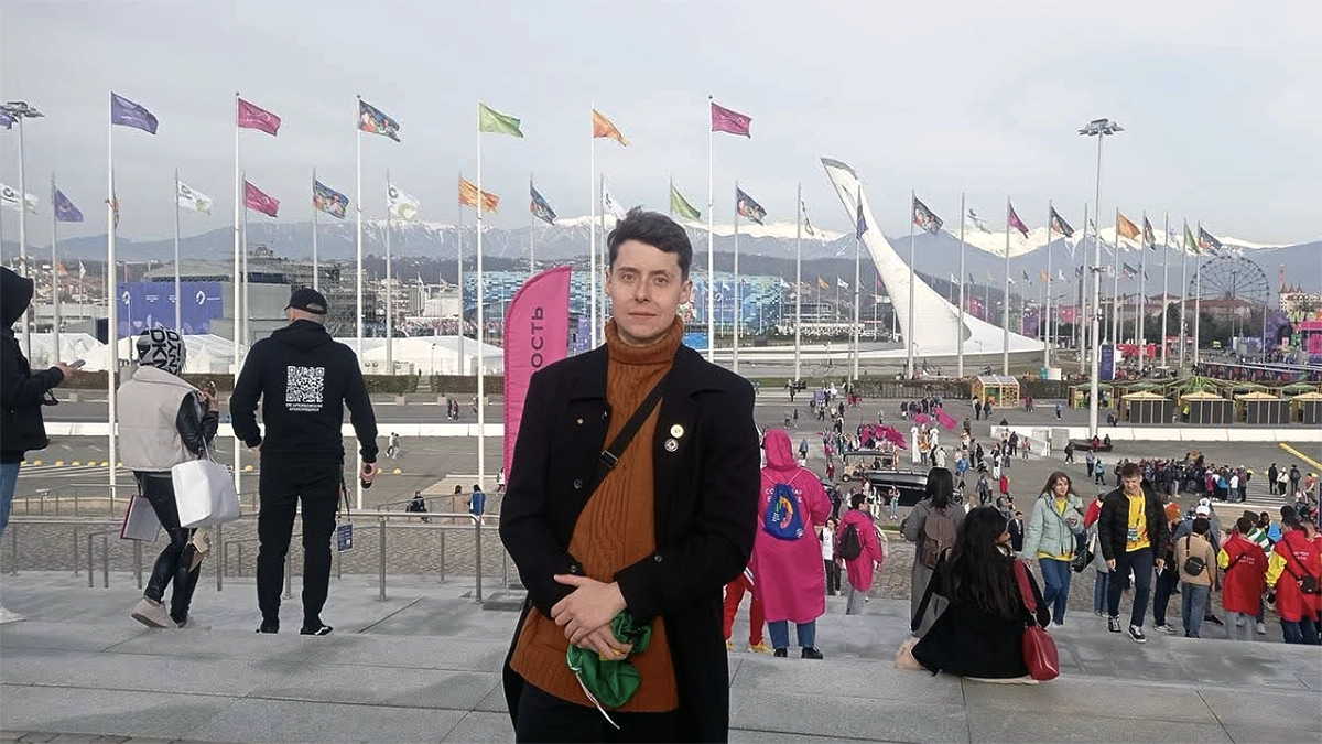 Jovem brasileiro posa para foto em escadaria de um grande evento internacional na Rússia, com bandeiras coloridas ao fundo e montanhas nevadas no horizonte. Ao redor, pessoas de diversas nacionalidades circulam pelo espaço, criando um ambiente vibrante e multicultural.