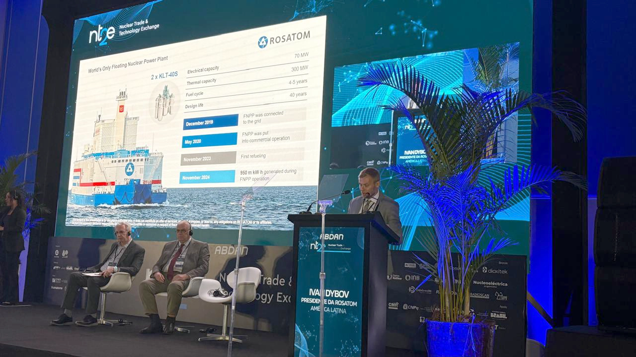 Ivan Dybov, presidente da Rosatom América Latina, discursa em um palco do fórum NT2E-2025, diante de um telão que exibe informações sobre a única usina nuclear flutuante do mundo. Ao lado do púlpito, dois palestrantes estão sentados. O ambiente é moderno, com iluminação azul e telão digital ao fundo.