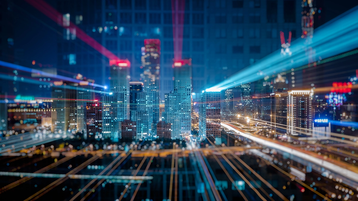 Imagem noturna de uma cidade moderna com vários arranha-céus iluminados. A foto apresenta um efeito visual de longa exposição com rastros de luz que se estendem pelas ruas e fachadas dos edifícios, criando uma sensação de velocidade e tecnologia. Os prédios ao centro têm luzes vermelhas no topo, e há feixes de luz azulados cruzando a imagem horizontalmente. A composição transmite uma atmosfera futurista e urbana.