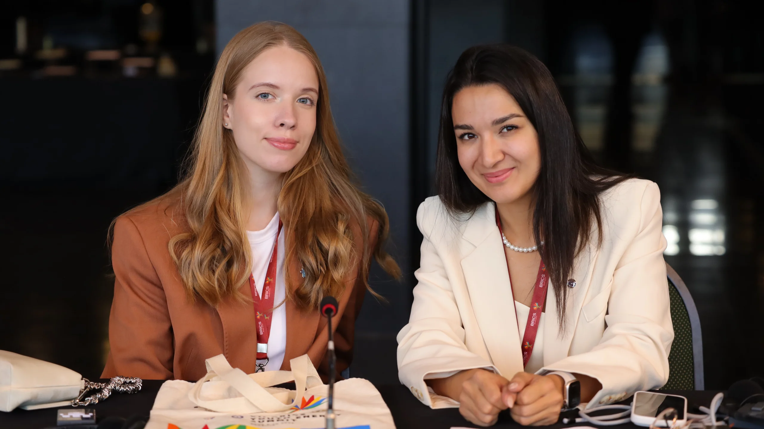 A ativista Altynay Bakhtina (à direita) e uma colega delegada estão sentadas lado a lado em uma mesa de conferência, ambas sorrindo. Elas usam crachás de identificação e estão posicionadas em frente a materiais do evento, como ecobag, celular, microfone e bloco de notas. A ecobag traz a marca do Global Youth Summit, indicando o contexto de participação juvenil em pautas globais. O fundo escuro e desfocado reforça o ambiente formal e institucional do evento.