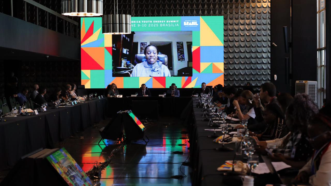 A imagem mostra uma reunião formal com uma longa mesa em formato retangular, onde jovens delegados dos países dos BRICS estão reunidos para a sessão da Plataforma Nuclear, realizada durante a BRICS Youth Energy Summit, nos dias 9 e 10 de junho de 2025, em Brasília. Ao fundo, destaca-se um telão colorido com identidade visual vibrante do evento, transmitindo a fala de uma participante remota. Em primeiro plano, é possível ver laptops, blocos de anotações, microfones e ecobags personalizadas com a marca do evento.