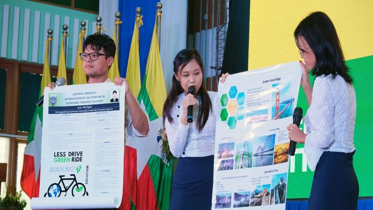 Três estudantes apresentam pôsteres sobre soluções de transporte sustentável e projetos de engenharia em um evento acadêmico, segurando microfones e explicando suas ideias ao público.