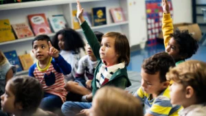 Crianças pequenas sentadas no chão de uma sala de aula, atentas e participativas. Algumas levantam a mão para responder a perguntas. Ao fundo, há estantes com livros infantis coloridos. A cena transmite um ambiente acolhedor e interativo de ensino na educação infantil.