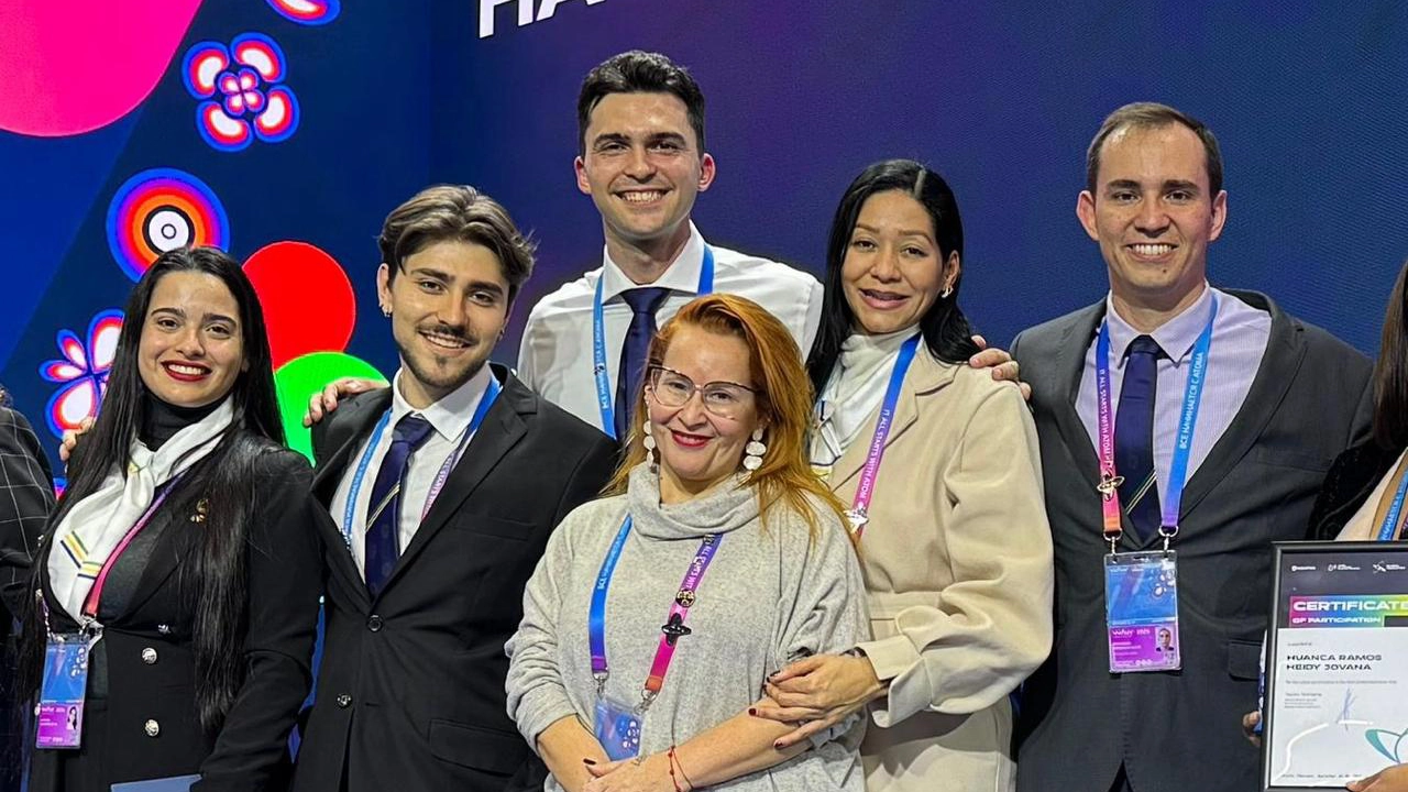 Seis pessoas estão em pé, lado a lado, sorrindo para a câmera em frente a um painel colorido com formas circulares e florais. Todas usam crachás pendurados no pescoço, indicando participação em um evento. Três homens estão de terno e gravata, e três mulheres usam roupas formais, incluindo um casaco bege, um vestido preto com lenço branco e uma blusa cinza clara com gola alta. Uma delas segura um certificado de participação. O grupo transmite alegria e espírito de equipe.