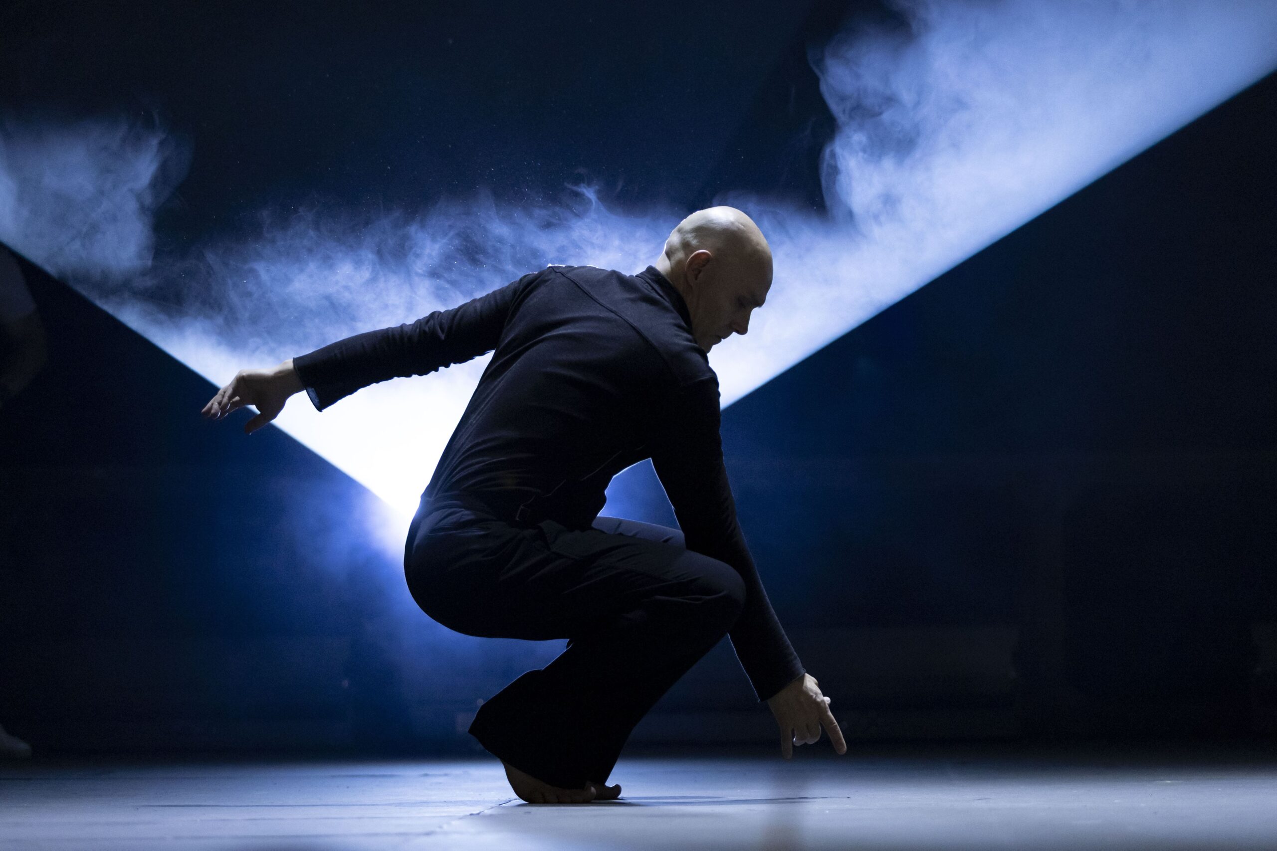 Bailarino vestido de preto, agachado no palco, com uma das mãos apontando para o chão e um feixe de luz diagonal iluminando fumaça atrás dele, criando atmosfera intensa e dramática