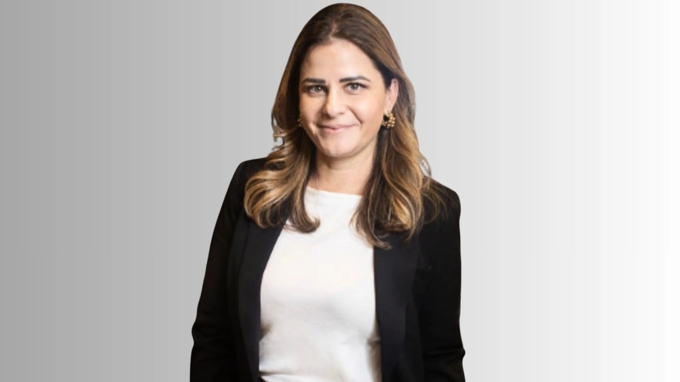 Retrato profissional, meio corpo, de uma mulher de cabelos castanhos na altura dos ombros, usando blazer preto e blusa branca, com leve sorriso e olhando para a câmera; fundo liso em degradê cinza, aparência corporativa