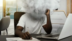 Homem sentado à mesa de trabalho com a cabeça substituída por uma nuvem densa, simbolizando o impacto da má qualidade do ar interno na saúde e no desempenho