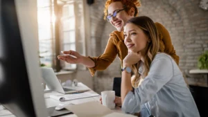 Duas mulheres trabalham juntas em um escritório. Uma delas, de cabelos ruivos e óculos, sorri enquanto aponta para a tela do computador. A outra, de cabelos longos e castanhos, observa atenta, apoiando o queixo na mão e segurando uma caneca branca.