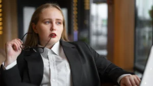 Mulher em traje formal preto e branco, sentada e segurando óculos junto ao rosto, com expressão séria e contemplativa em ambiente corporativo