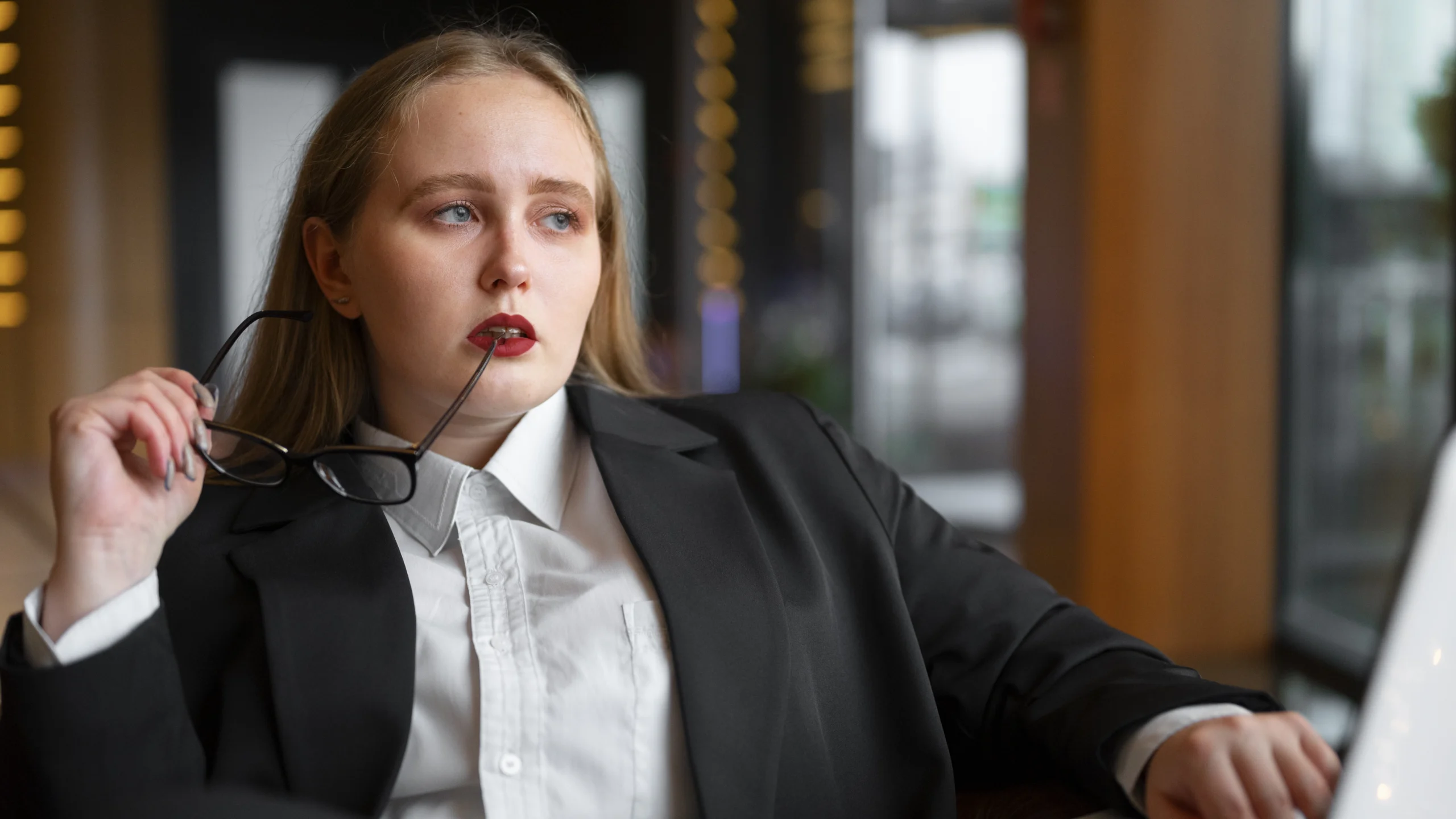 Mulher em traje formal preto e branco, sentada e segurando óculos junto ao rosto, com expressão séria e contemplativa em ambiente corporativo