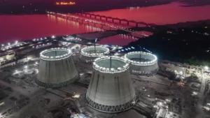 Foto aérea noturna de um canteiro industrial com várias torres de resfriamento circulares em construção, iluminadas por refletores. Ao fundo, um grande rio com uma ponte cheia de luzes e o céu com tom arroxeado.