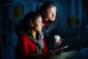Uma mulher olha para a tela de um computador enquanto segura um tablet. Ao lado dela, um homem se inclina para observar o monitor, segurando um copo descartável. Ambos parecem concentrados e trabalham em um escritório com iluminação baixa