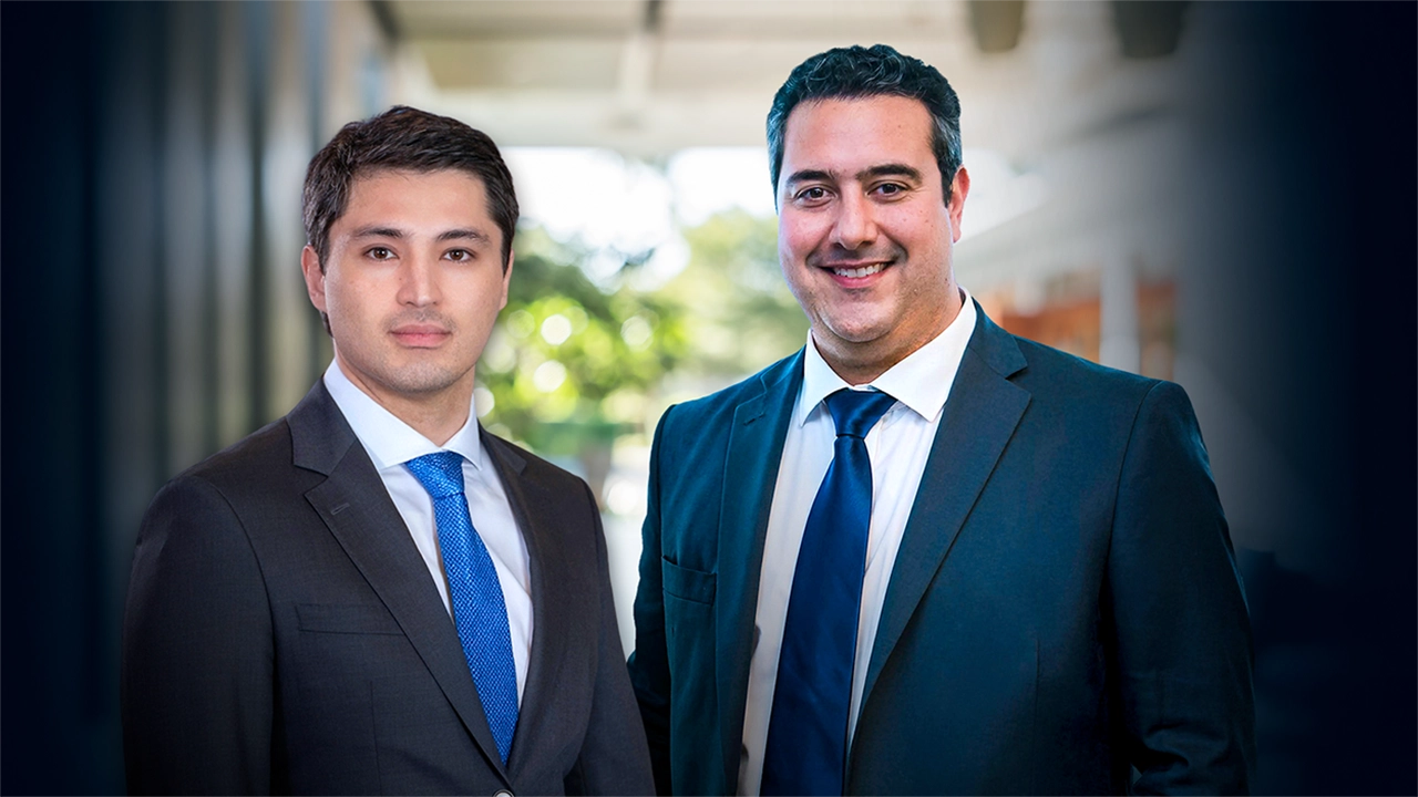 Dois homens de terno em ambiente corporativo: um à esquerda com terno cinza e gravata azul-clara, e outro à direita com terno escuro e gravata azul-marinho, sorrindo, com fundo desfocado.