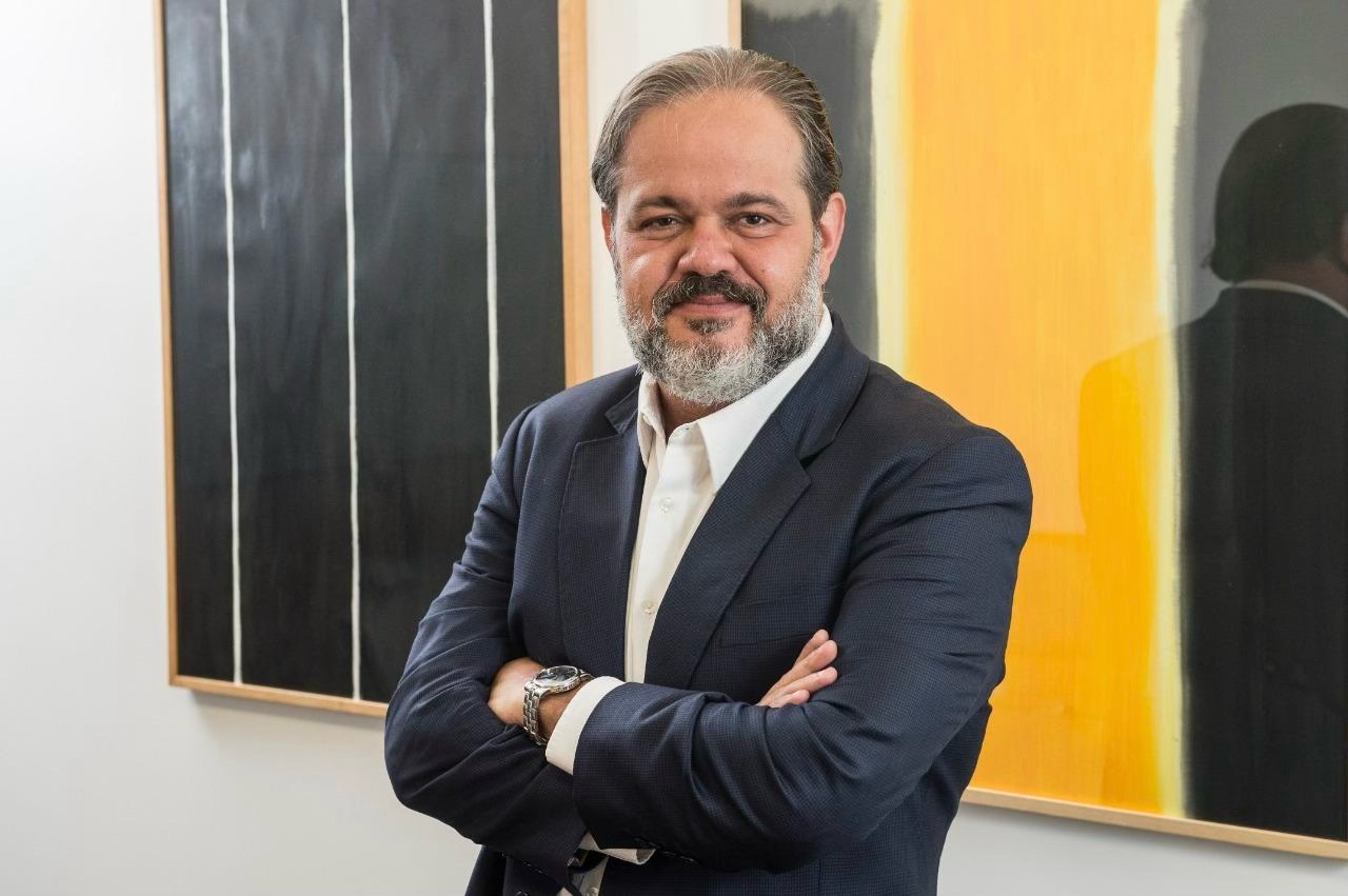 Homem de meia-idade, com cabelos grisalhos penteados para trás e barba curta também grisalha, veste terno azul-marinho e camisa branca. Está de braços cruzados, usando relógio metálico no pulso esquerdo, e olha para a câmera com expressão confiante. Ao fundo, há quadros abstratos em preto, branco e amarelo pendurados na parede clara.