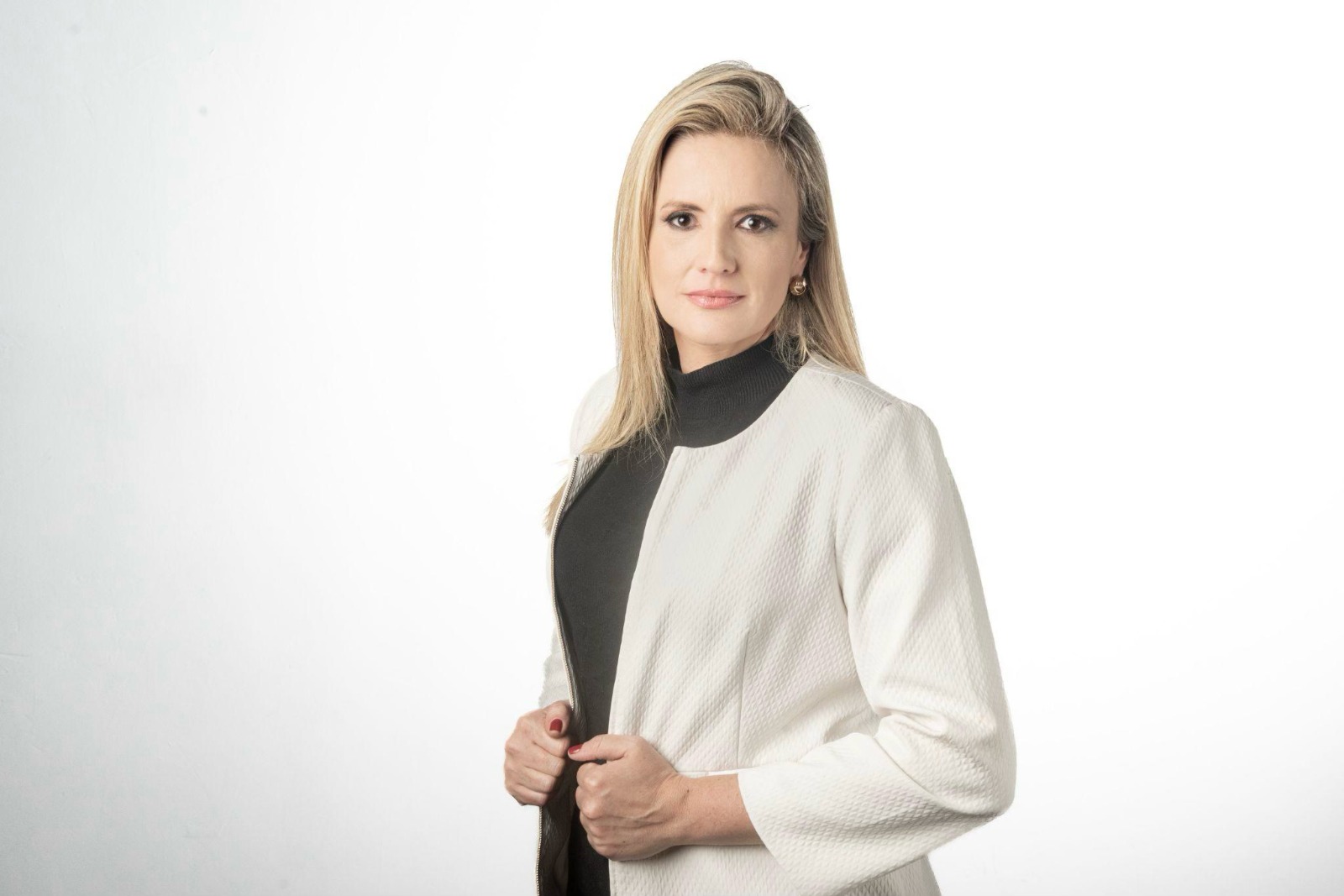 Mulher de pele clara e cabelos loiros lisos, na altura dos ombros, posa de frente para a câmera com expressão séria e postura ereta. Veste blusa preta de gola alta e casaco claro de textura discreta, segurando a lapela com as duas mãos. Usa brincos pequenos e esmalte vermelho. O fundo é neutro e claro, com iluminação suave e uniforme.
