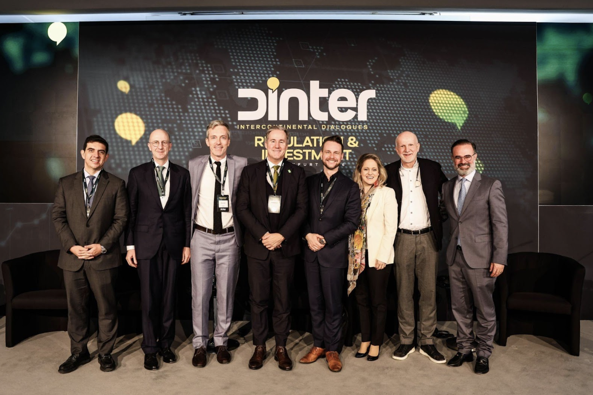 Grupo de participantes do evento Regulation & Investment – Frankfurt 2026 posa para foto no palco, diante de painel com a marca do Dinter – Intercontinental Dialogues. Os especialistas participaram de debate sobre financiamento, regulação e cadeias industriais da economia circular no setor de energia verde.