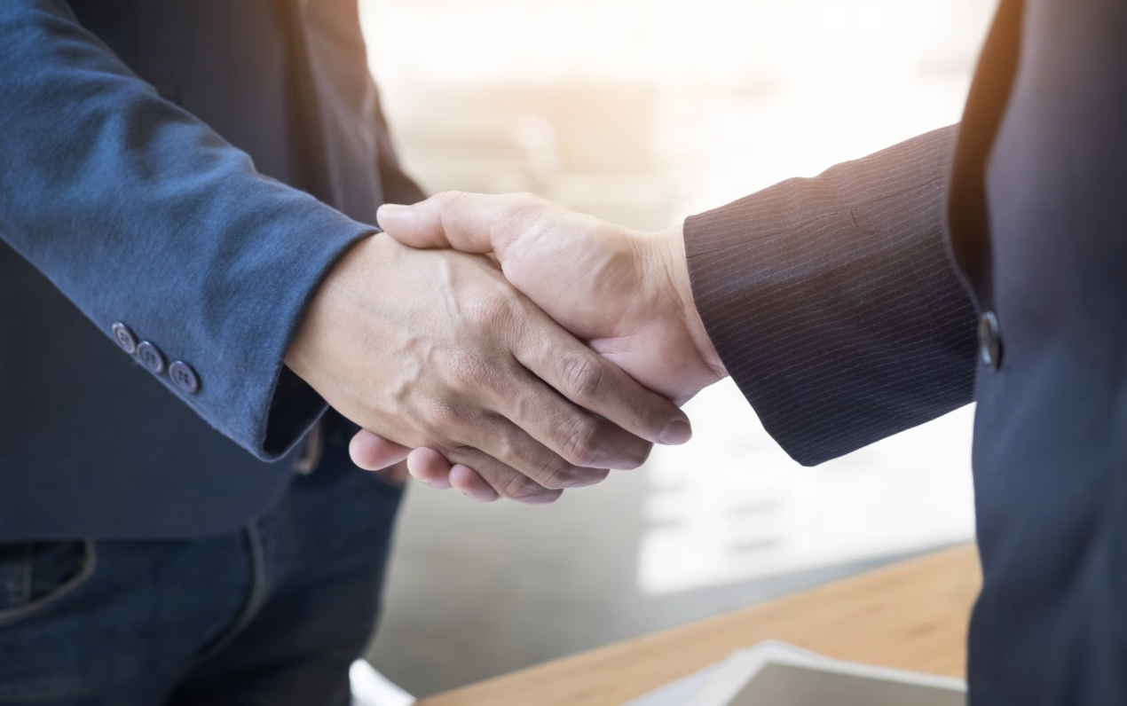 Duas pessoas de terno apertam as mãos em um aperto firme, em ambiente corporativo, com mesa e documentos ao fundo.