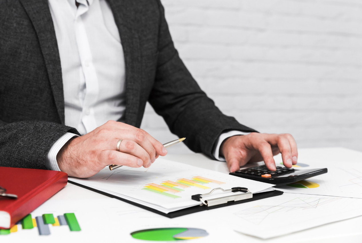 Pessoa de terno analisa gráficos financeiros em prancheta enquanto utiliza uma calculadora sobre a mesa, com relatórios e indicadores coloridos ao redor.