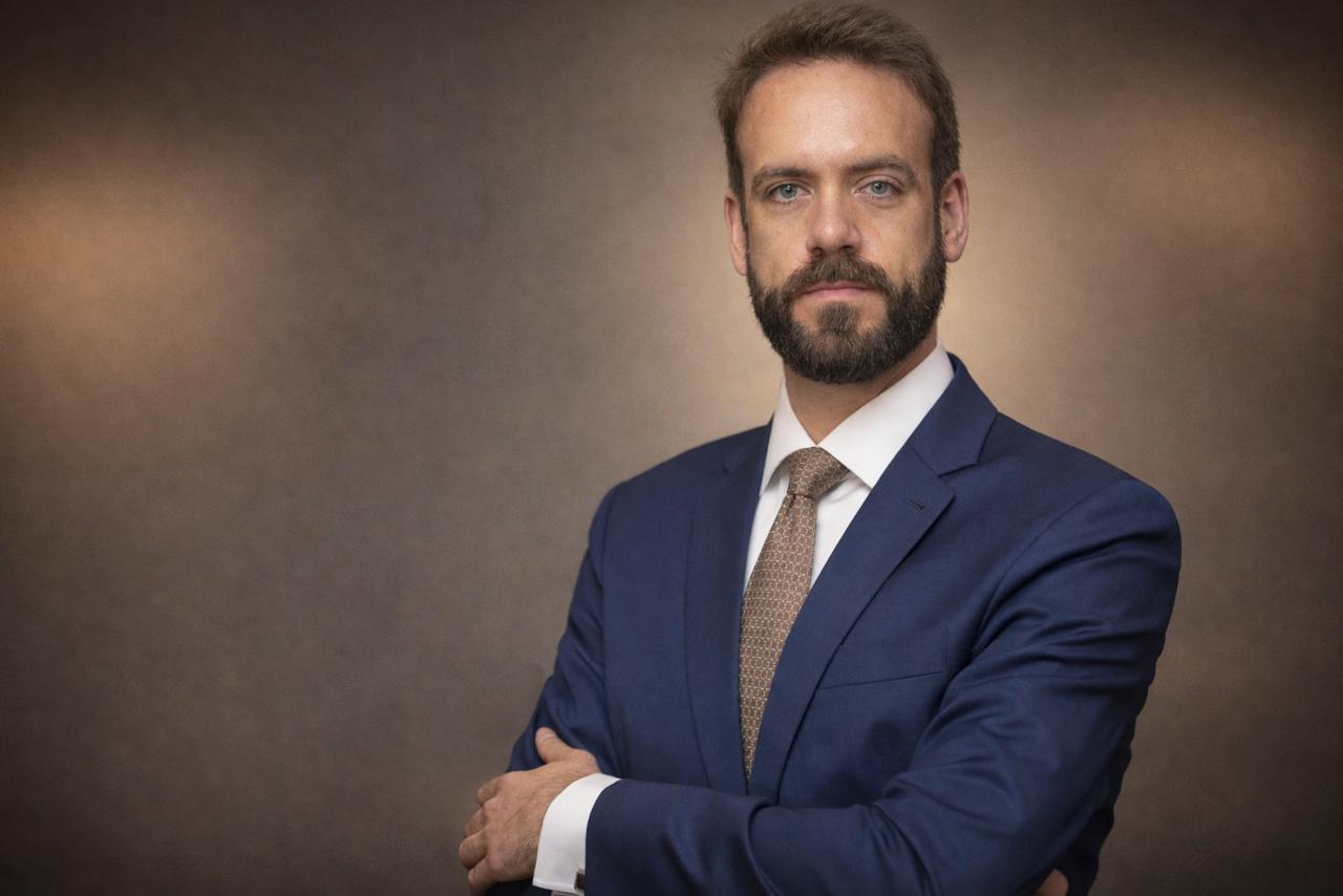 Homem adulto de pele clara posa para retrato formal em estúdio. Ele tem cabelo castanho curto e barba cheia bem aparada. Veste terno azul, camisa branca e gravata bege. Está com os braços cruzados e expressão séria, olhando diretamente para a câmera. O fundo é neutro, em tons suaves de marrom desfocado, típico de fotografia corporativa ou institucional. A iluminação é suave e frontal, destacando o rosto e o traje.