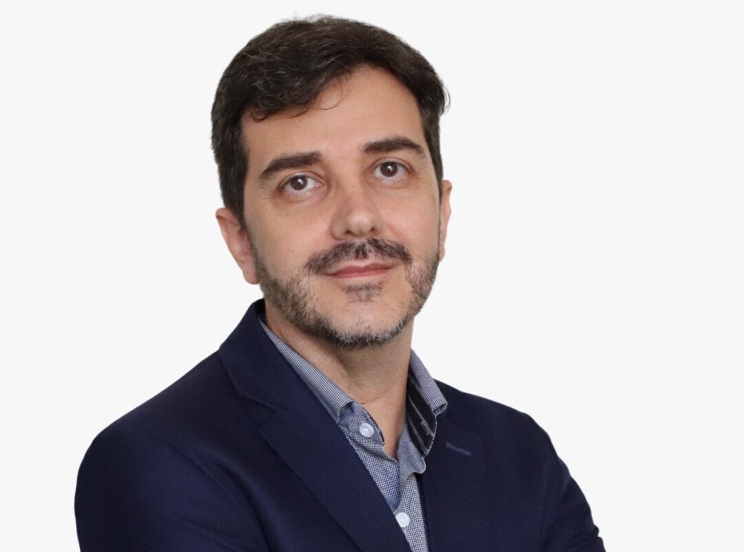 Homem de cabelo escuro e barba curta, usando paletó azul-marinho e camisa azul-clara, posa de braços cruzados diante de fundo cinza claro. Ele olha para a câmera com expressão serena e usa relógio no pulso e aliança na mão esquerda.