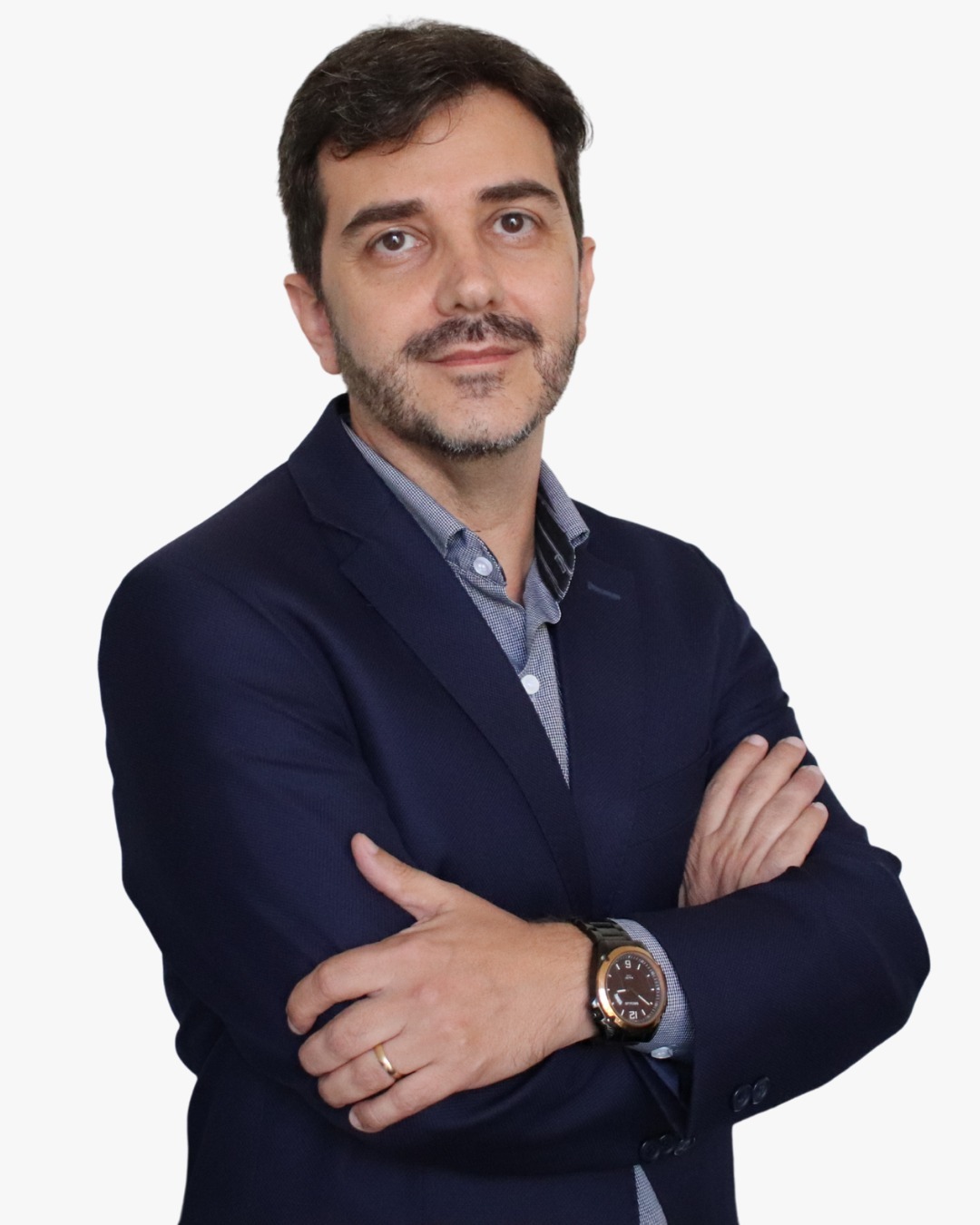 Homem de cabelo escuro e barba curta, usando paletó azul-marinho e camisa azul-clara, posa de braços cruzados diante de fundo cinza claro. Ele olha para a câmera com expressão serena e usa relógio no pulso e aliança na mão esquerda.