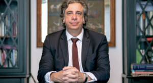 Homem de meia-idade, vestindo terno escuro e gravata vinho, sentado com as mãos entrelaçadas à frente, em ambiente interno com estante de livros e quadro ao fundo.