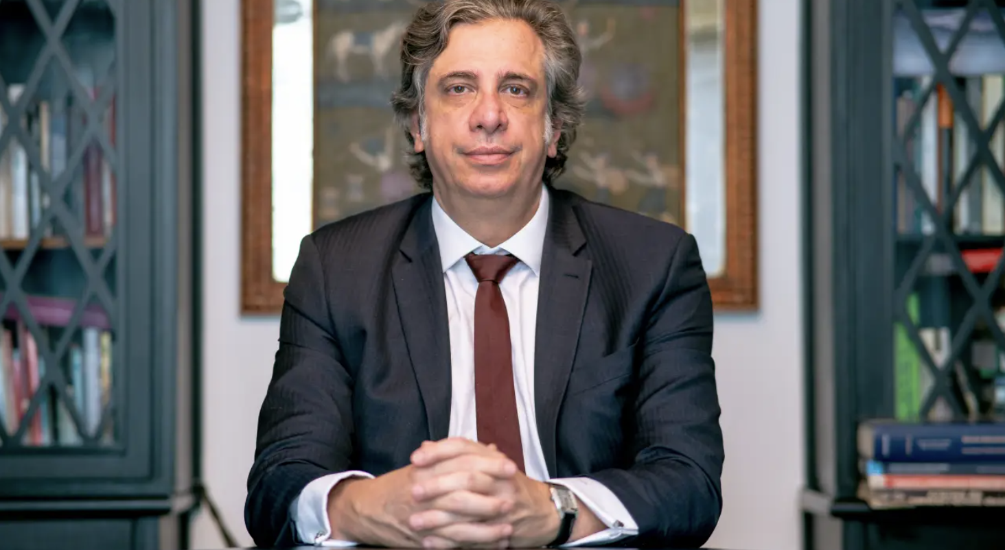 Homem de meia-idade, vestindo terno escuro e gravata vinho, sentado com as mãos entrelaçadas à frente, em ambiente interno com estante de livros e quadro ao fundo.