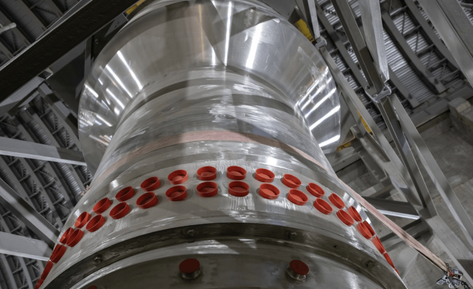 Grande equipamento cilíndrico de metal em instalação industrial, com fileiras de aberturas vedadas por tampas vermelhas e estrutura de aço ao redor.