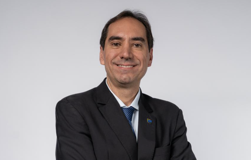 retrato em estúdio de homem de terno escuro, camisa branca e gravata azul, sorrindo de forma discreta, com os braços cruzados, relógio no pulso e fundo cinza-claro.