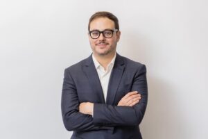 Homem de terno azul-marinho e camisa branca, usando óculos, posa de braços cruzados diante de fundo claro. Ele olha para a câmera com expressão leve e postura formal, em retrato corporativo de estúdio.