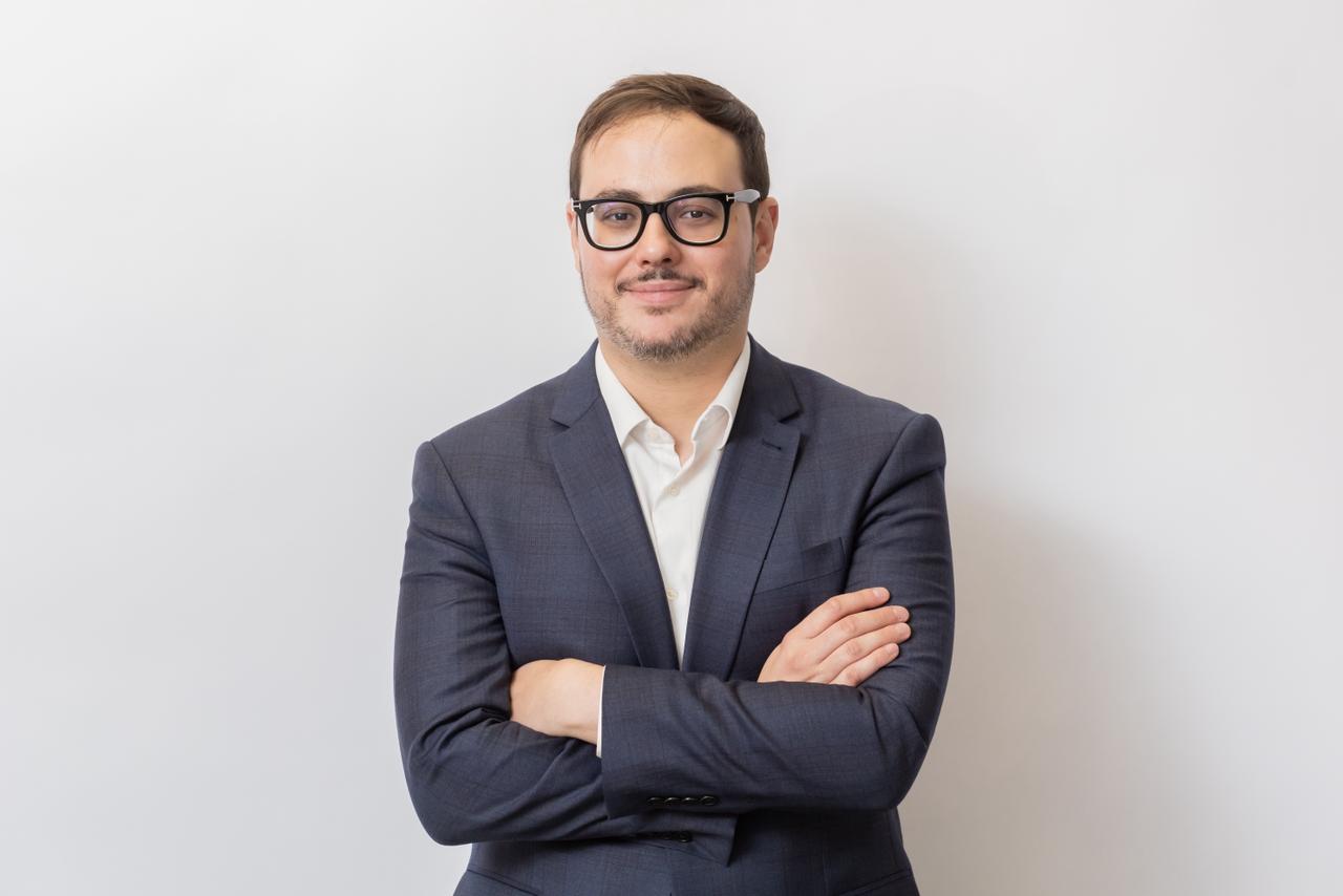 Homem de terno azul-marinho e camisa branca, usando óculos, posa de braços cruzados diante de fundo claro. Ele olha para a câmera com expressão leve e postura formal, em retrato corporativo de estúdio.