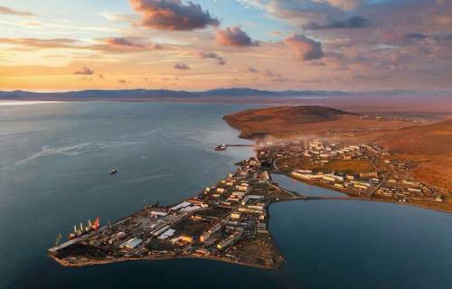Vista aérea de porto costeiro no Ártico, com navios atracados, áreas industriais e estruturas logísticas próximas a uma pequena cidade, às margens de uma baía sob céu alaranjado.