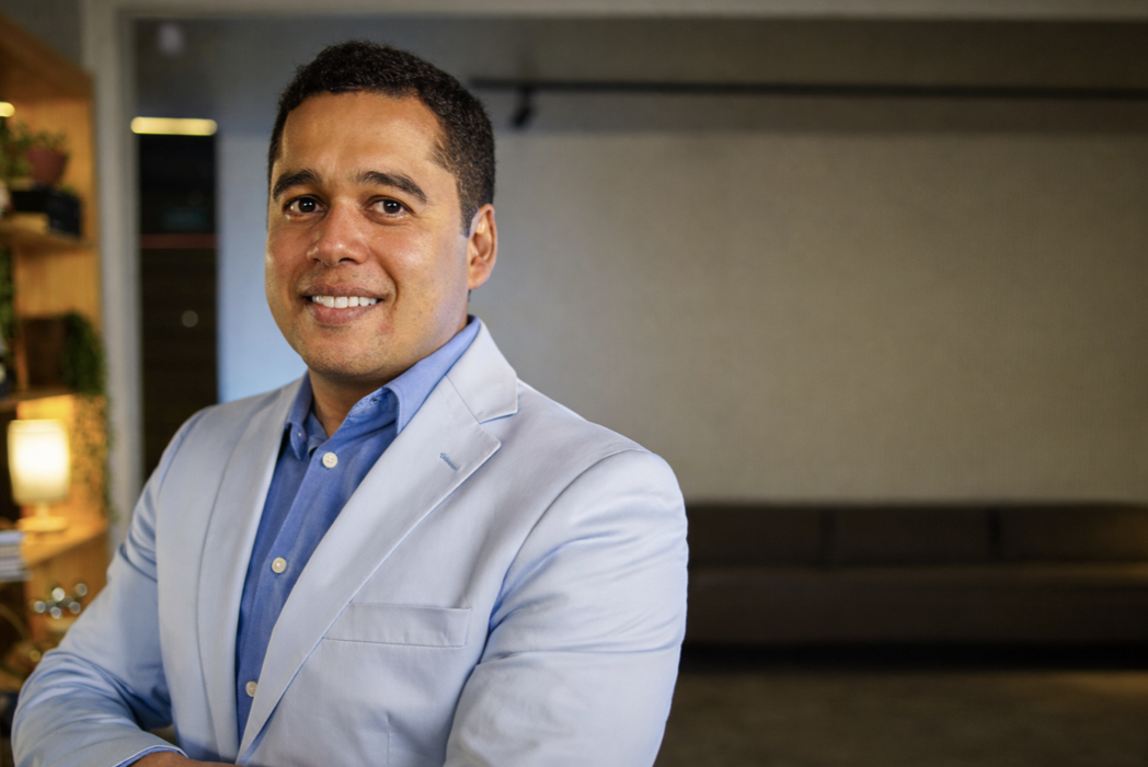 Homem sorridente posa com os braços cruzados em ambiente interno, usando blazer azul-claro e camisa azul.