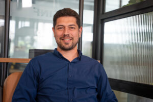 Homem de camisa azul sentado em ambiente corporativo, sorrindo para a câmera, com divisórias de vidro ao fundo.