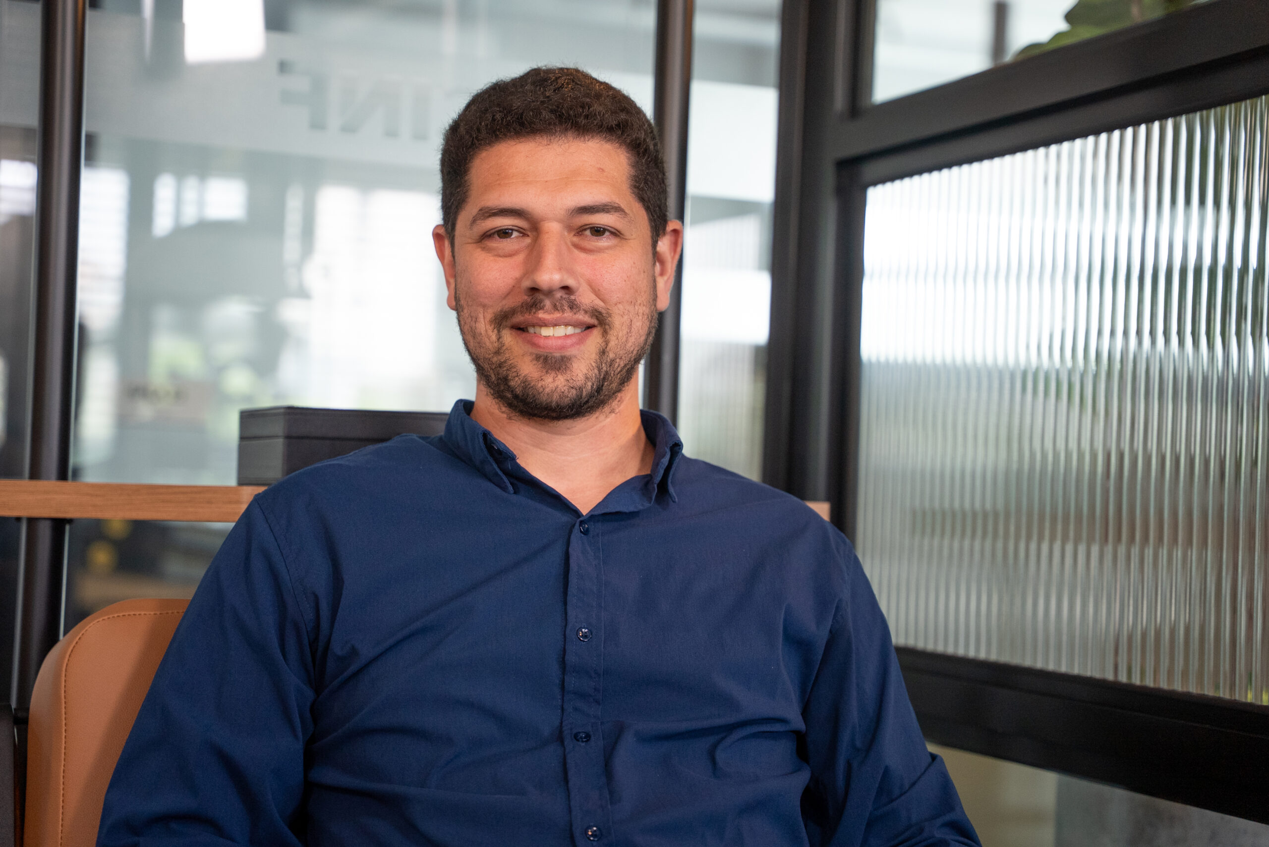 Homem de camisa azul sentado em ambiente corporativo, sorrindo para a câmera, com divisórias de vidro ao fundo.