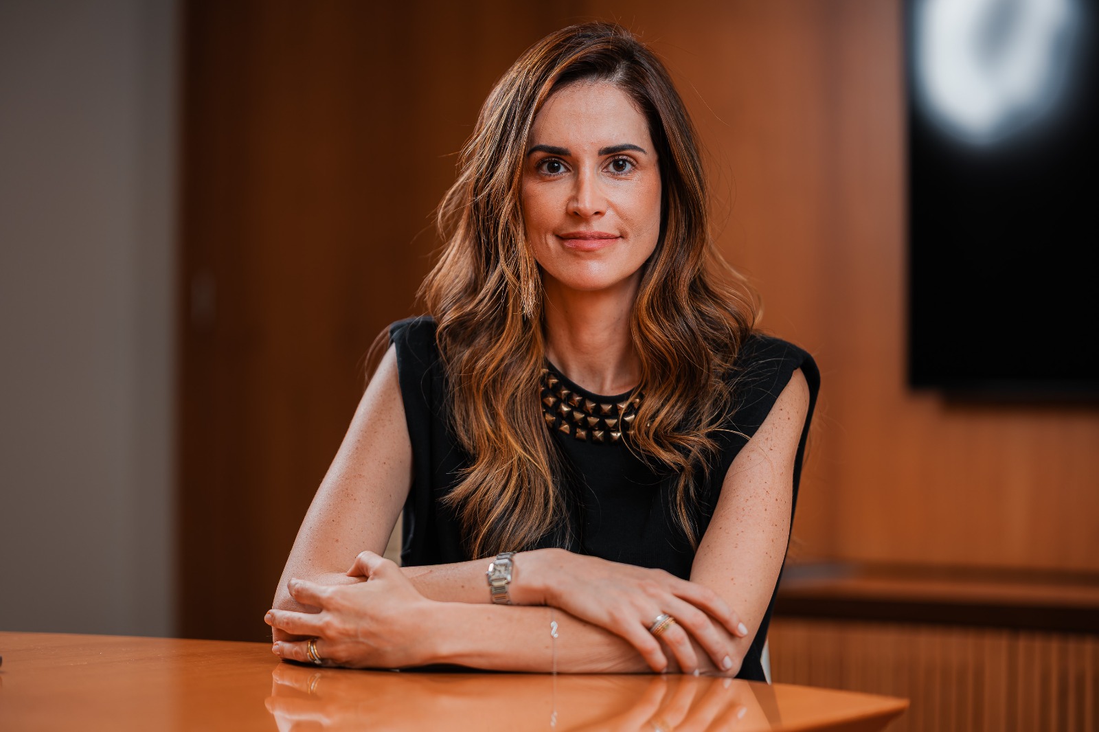 Mulher de cabelos longos posa sentada à mesa em ambiente corporativo, usando blusa preta e olhando para a câmera.