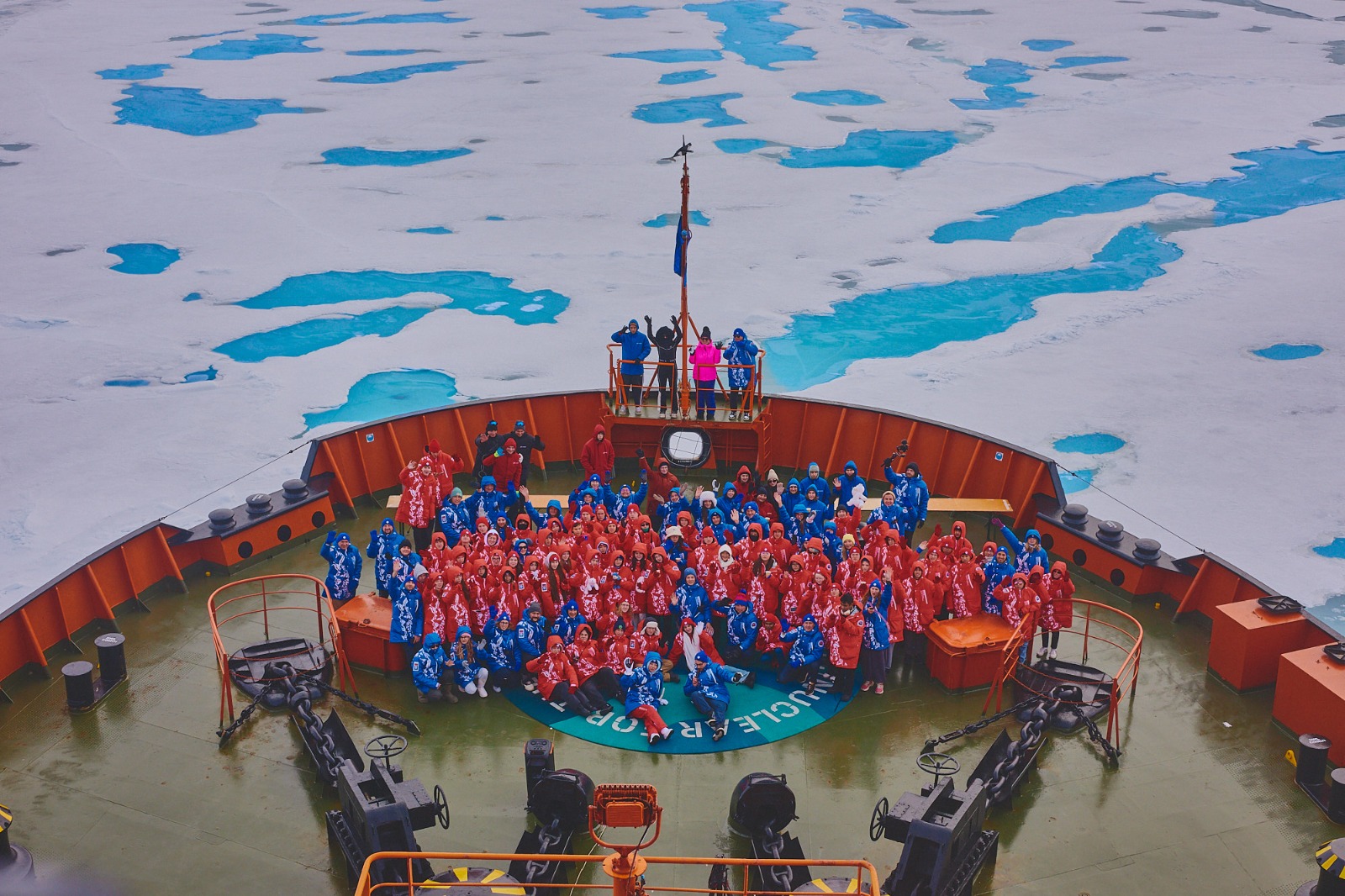 Grupo de pessoas com roupas térmicas vermelhas e azuis posa na proa de um navio cercado por gelo e águas azuladas em região polar.