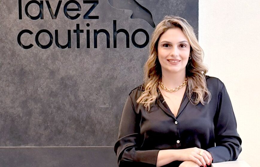 Mulher posa em ambiente corporativo, ao lado de parede com o logotipo do escritório Lavez Coutinho, vestindo camisa preta, saia marrom e sapatos de salto