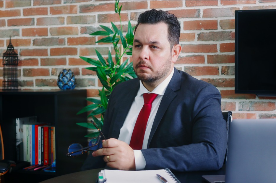 Homem de terno escuro e gravata vermelha aparece sentado em ambiente de escritório, segurando óculos, com notebook, caderno e livros ao fundo, diante de uma parede de tijolos aparentes.
