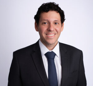 Homem em traje formal posa sorrindo para retrato profissional em fundo claro.
