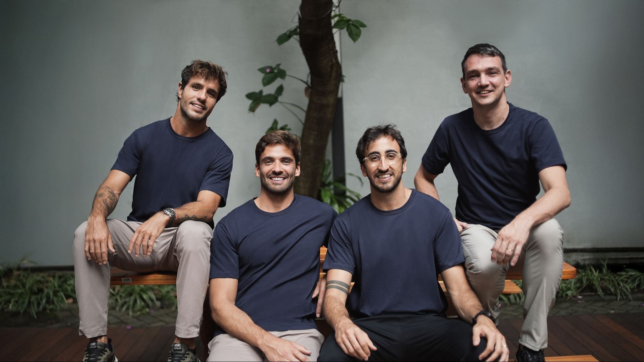**Texto alternativo** Quatro homens posam sorrindo em área externa com jardim ao fundo, usando camisetas azul-marinho e calças claras, sentados e apoiados em bancos de madeira.