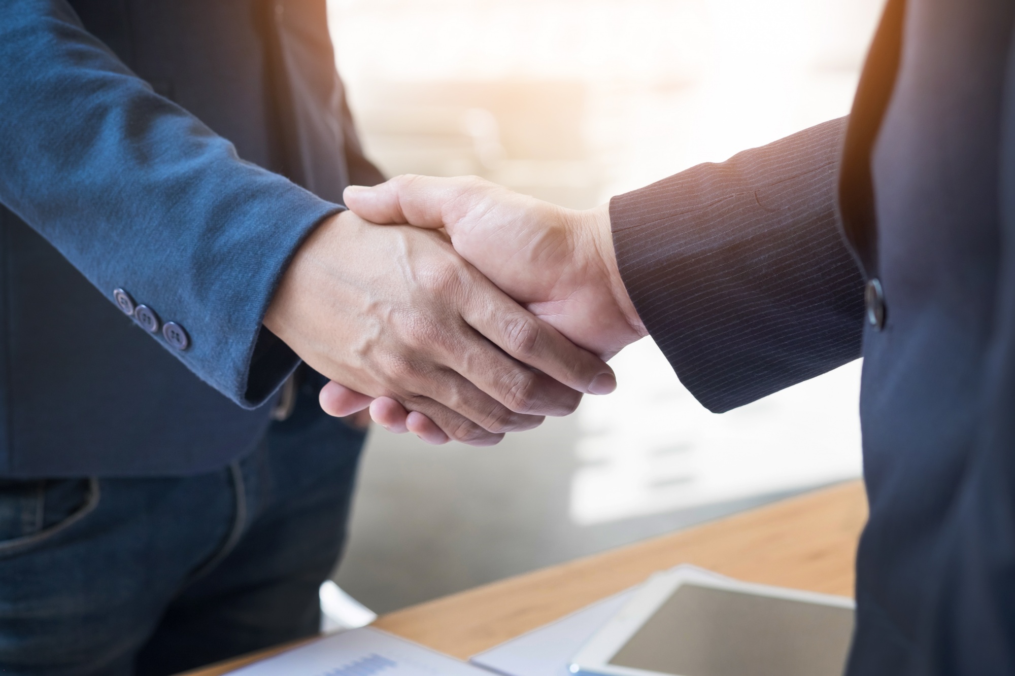 Duas pessoas de terno apertam as mãos em ambiente corporativo, simbolizando acordo, parceria ou fechamento de negócio.