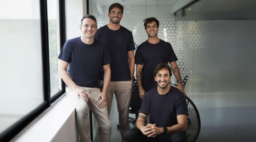 Quatro fundadores da Inspira posam sorrindo em um escritório moderno, com paredes de vidro ao fundo. Três estão em pé e um está sentado à frente, todos usando camisetas escuras