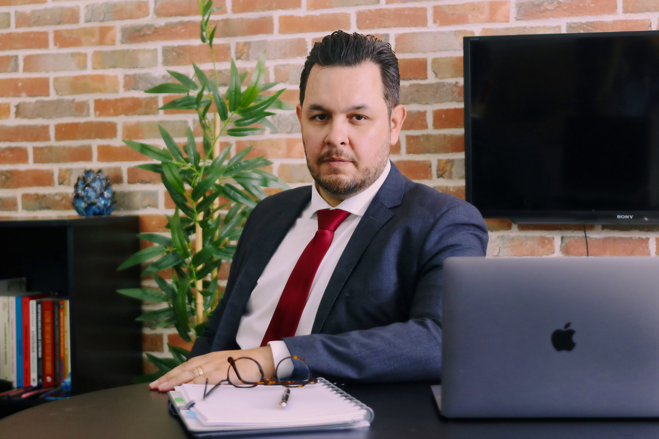 Retrato corporativo de um homem de terno azul e gravata vermelha sentado em um escritório, com notebook, caderno e óculos sobre a mesa. Ao fundo, há uma parede de tijolos, planta, estante de livros e uma televisão