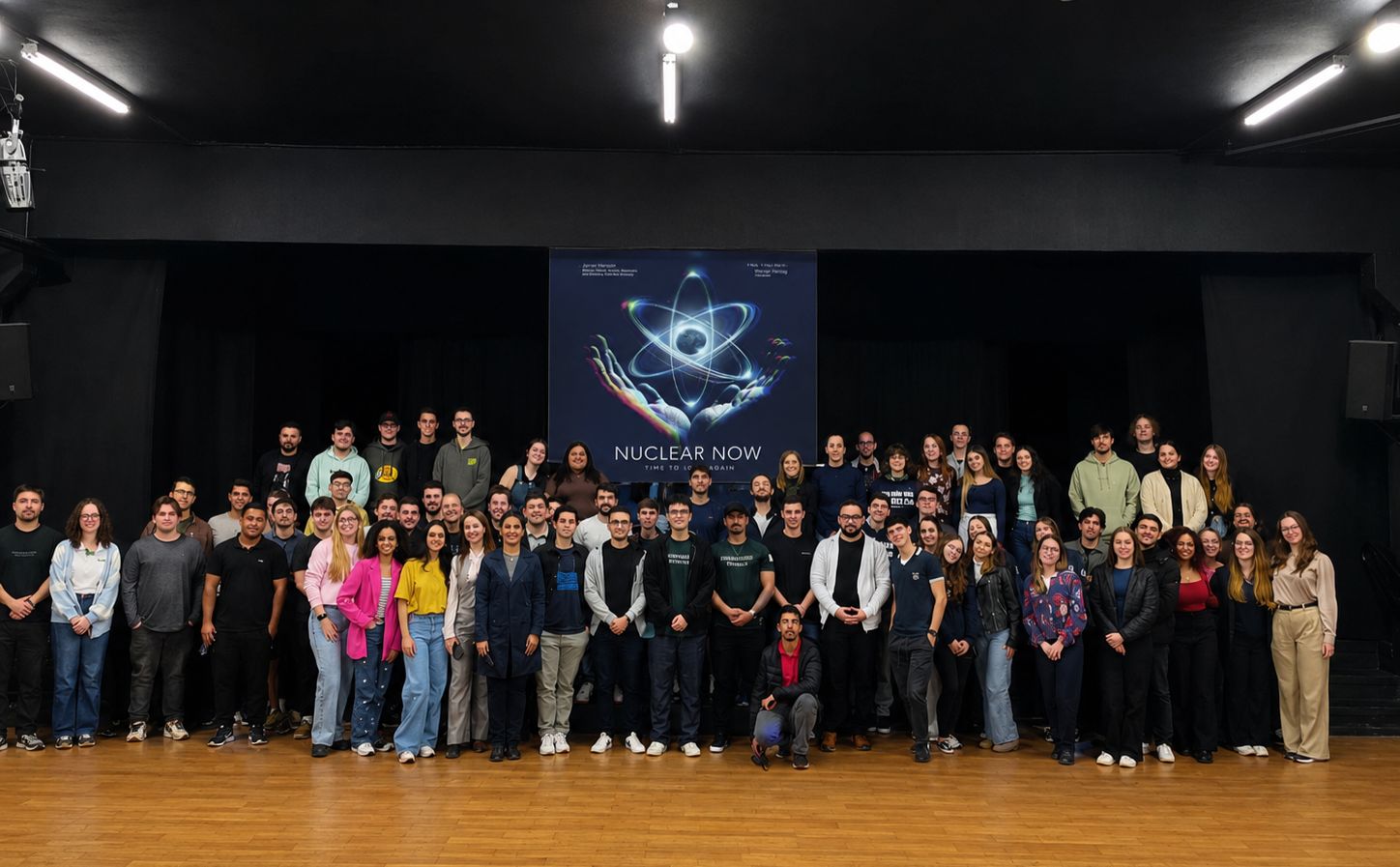 Grupo numeroso posa em um auditório, diante de um cartaz do documentário Nuclear Now. A imagem mostra participantes reunidos após sessão sobre energia nuclear, com palco, piso de madeira e fundo escuro.
