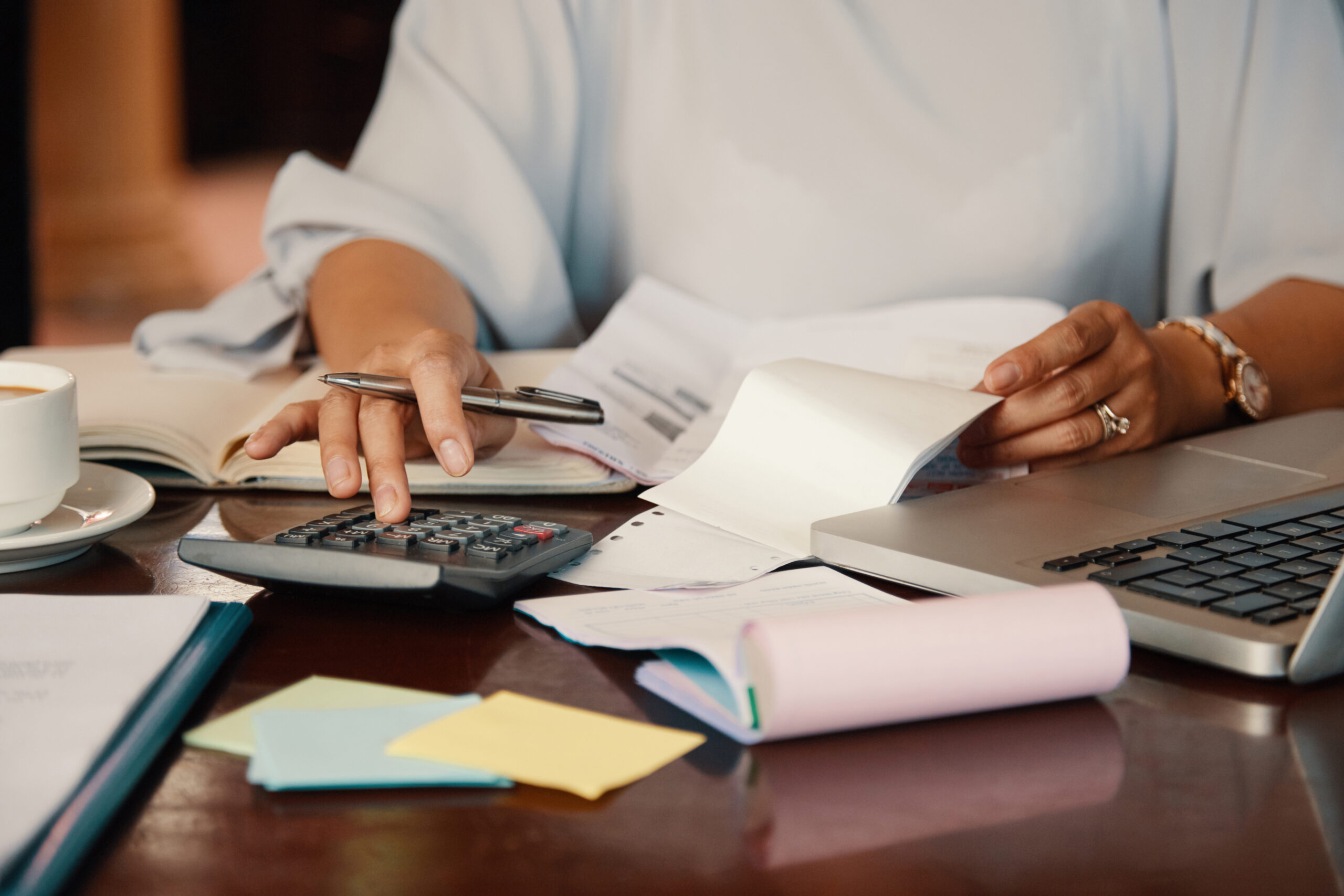 Pessoa confere documentos e usa calculadora ao lado de notebook, representando revisão financeira, controle de custos e análise tributária