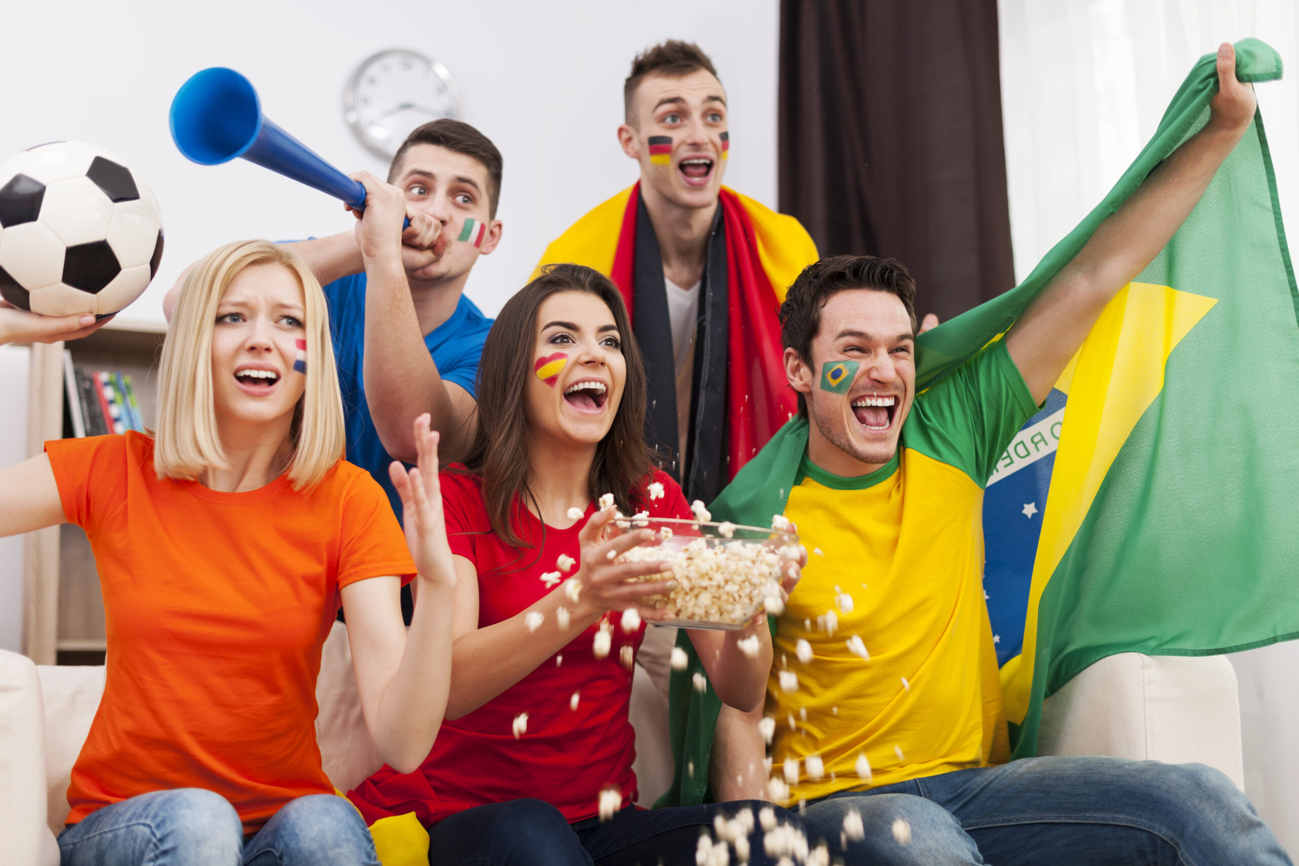 Grupo de torcedores assiste a uma partida de futebol em uma sala, com bandeira do Brasil, corneta, bola e pipoca, celebrando com entusiasmo